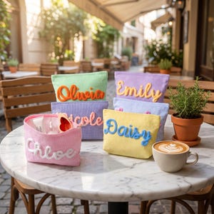 May include: A collection of colorful corduroy cosmetic bags, each with a name embroidered on the front. The bags are in various colors, including pink, green, purple, blue, and yellow. A cup of coffee and a potted plant are also on the table.