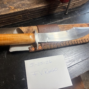 Puede incluir: Un cuchillo de hoja fija con una hoja de metal plateado pulido y un mango de madera de color marrón claro liso. El cuchillo descansa sobre una funda de cuero marrón con acabado texturizado y bordes cosidos, junto a una regla metálica en centímetros.