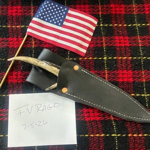 May include: A knife with a curved antler handle inside a black leather sheath with white stitching and two copper rivets, accompanied by a small American flag on a wooden stick and a white paper note with the handwritten text F V RAGO 7-5-26.