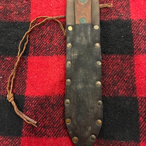 May include: A fixed-blade knife with a brown wooden handle secured by two green rivets, housed in a dark brown leather sheath. The sheath features a row of brass-colored metal rivets along both edges and includes a thin leather cord attached to the side.