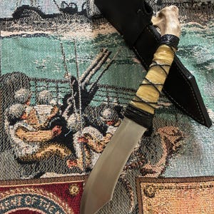 May include: A fixed-blade knife with a metallic silver blade and a handle crafted from a light-colored bone. The handle is wrapped in thin black cord and is positioned next to a black leather sheath with visible stitching along the edges.