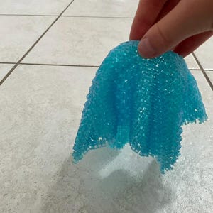 Puede incluir: Una almohadilla azul, texturizada y flexible es sostenida por una mano. La almohadilla está hecha de pequeñas estructuras interconectadas en forma de cuentas. La almohadilla es de un color azul brillante y vibrante. El fondo es un suelo de baldosas de color claro.