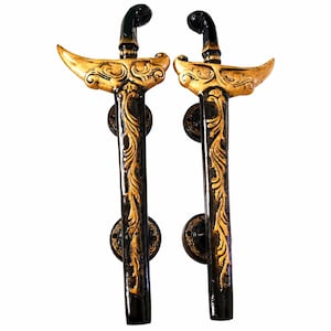Op de afbeelding: Twee decoratieve deurklinken in de vorm van traditionele Indonesische Kris-dolken. De handgrepen hebben een zwart-gouden kleurenschema met sierlijke, wervelende patronen. De handgrepen zijn gemonteerd op ronde, donkergekleurde achterplaten. Ongeveer 30 cm lang.
