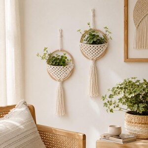 May include: Two wall-mounted macrame plant hangers featuring circular wooden hoops and off-white knotted cord pockets. Each hanger includes a long, dangling tassel at the base and holds a small green plant, suspended by a matching cord loop.