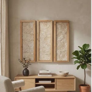 Op de afbeelding: Drie rechthoekige wandkunstwerken met licht houten frames. Elk stuk heeft een textuur, gebroken wit ontwerp. De kunst is gemonteerd op een neutraal gekleurde muur boven een houten kast. Een kleine plant en decoratieve items staan op de kast.