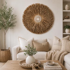 May include: A circular wall decoration made of natural fibres, resembling a sunburst, hangs above a cream sofa with cushions and a knitted throw. A wooden coffee table displays a vase, wooden beads, and books.