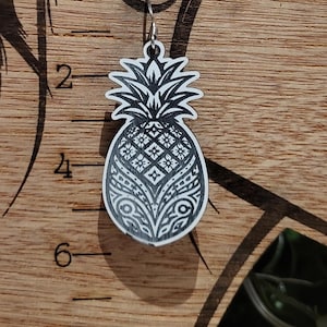 May include: A single silver-tone earring with a white and black pineapple design. The pineapple has a detailed pattern. The earring is hanging on a wooden display with a woman's face etched into it.