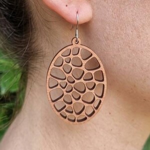 Peut inclure: Boucles d'oreilles ovales en bois clair, avec un motif découpé de formes irrégulières. Les boucles d'oreilles sont de couleur marron clair et sont suspendues à des crochets argentés.