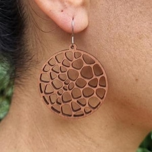 Peut inclure: Gros plan sur une boucle d'oreille ronde en bois marron avec un motif ajouré détaillé. La boucle d'oreille est suspendue à un crochet argenté. Le motif présente un réseau de formes interconnectées. La boucle d'oreille est portée à l'oreille.
