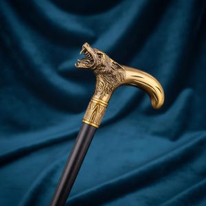 Puede incluir: Un bastón con un mango de metal dorado en forma de cabeza de lobo gruñendo, con detalles de pelaje y dientes visibles. El mango se conecta a un eje negro liso y recto mediante un collar decorativo de color dorado con líneas verticales.