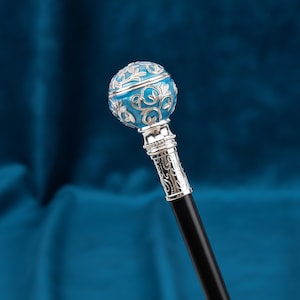 May include: A walking stick featuring a spherical handle with a bright blue enamel finish and intricate silver-toned floral filigree overlay. The handle connects to a smooth black shaft via a decorative silver-toned collar with embossed patterns.