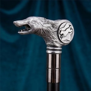 May include: A silver-toned metal walking stick handle shaped like a wolf head with an open mouth and detailed fur texture. A circular emblem on the side features a relief of a running wolf. The handle connects to a smooth, dark cylindrical shaft.