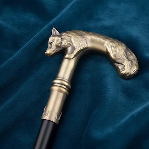 May include: A walking cane featuring a metallic bronze-toned handle sculpted in the shape of a wolf. The handle connects to a smooth black shaft via a decorative metallic collar with engraved detailing. The wolf figure shows detailed fur texture.