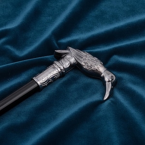 May include: A walking cane featuring a metallic silver-toned handle shaped like a bird head with detailed feather engravings. The handle connects to a smooth black shaft via a decorative silver-toned collar with intricate scrollwork patterns.