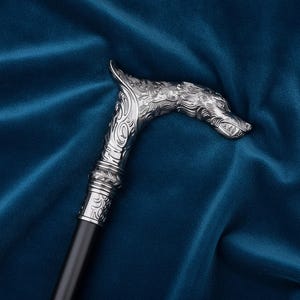 Könnte beinhalten: Ein Gehstock mit einem silberfarbenen Metallgriff in Form eines Wolfskopfes, der mit detaillierten Fellstrukturen und geschwungenen Mustern verziert ist. Der Griff ist mit einem glatten, geraden schwarzen Schaft verbunden, was einen Kontrast zwischen dem metallischen Oberteil und dem dunklen, einfarbigen unteren Teil des Stocks erzeugt.