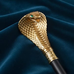 Può includere: Un'impugnatura per bastone da passeggio a forma di cobra dorato con un motivo a scaglie testurizzato e una piccola gemma verde incastonata nell'occhio. La testa del cobra è fissata a un fusto nero liscio con un collare decorativo in metallo dorato.