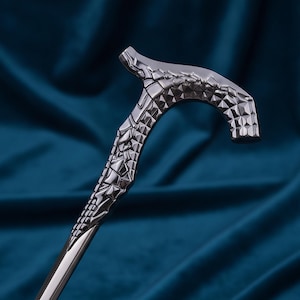 Könnte beinhalten: Ein metallischer Gehstockgriff mit dunkler, polierter Oberfläche und einer komplexen, facettierten geometrischen Textur, die an gesprungenen Stein oder Schuppen erinnert. Der Griff hat eine geschwungene, ergonomische Form.