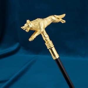 May include: A walking stick featuring a gold-colored handle shaped like a leaping wolf with an open mouth and detailed fur texture. The handle sits atop a smooth, straight, black shaft, with a decorative gold-colored collar connecting the two parts.
