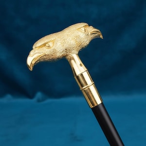 May include: A walking stick featuring a gold-colored handle shaped like a double-headed eagle. The eagle heads are detailed with textured feathers and sharp beaks. The handle connects to a smooth, straight black shaft with a gold-colored metal collar.