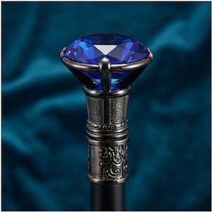 Puede incluir: Un mango de bastón con una gran piedra preciosa azul profundo facetada en un engaste de metal plateado. La base metálica debajo de la piedra tiene una superficie texturizada y grabada con patrones intrincados y un acabado oscuro.
