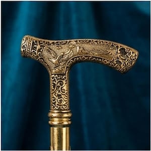 May include: An ornate gold-coloured walking cane with a T-shaped handle. The handle features an engraved bird design and decorative scrollwork. The shaft is a matching gold colour, and the cane appears to be made of metal.