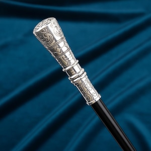 May include: A walking stick featuring a silver-toned handle with intricate embossed scrollwork and geometric patterns. The handle has a cylindrical shape with a rounded top, transitioning into a smooth, solid black shaft that extends downward.