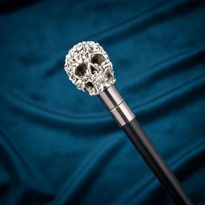 May include: A walking stick featuring a silver-toned skull handle with intricate, textured detailing. The skull is mounted on a smooth, dark black shaft, connected by a polished metallic collar that transitions between the handle and the main body.