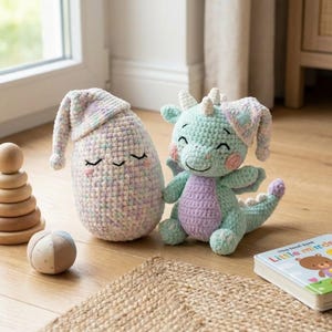May include: Two crocheted plush toys: a light green dragon with purple accents and a sleeping cap, and a pastel-colored egg-shaped figure wearing a matching cap. The toys are on a wooden floor, with a small book visible.