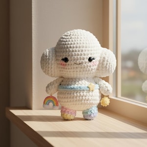 Puede incluir: Una muñeca de nube de ganchillo hecha a mano con una cara sonriente, mejillas rosadas y ojos negros. La nube lleva una banda azul claro con una estrella dorada y sostiene un arcoíris y una estrella amarilla. Los pies de la muñeca son de colores pastel arcoíris.