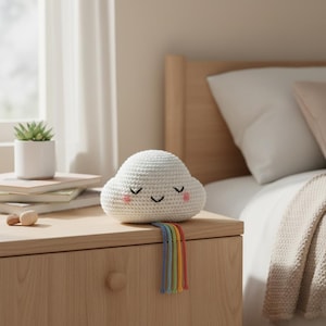 May include: A white crocheted cloud-shaped decorative item with a smiling face and pink cheeks. A rainbow of colorful yarn strands hangs from the cloud. The item is placed on a wooden nightstand next to a bed.