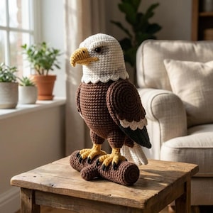 May include: A handcrafted crochet eagle figurine. The eagle has a white head, a yellow beak, and brown body with white and black accents on the wings. It is perched on a brown log with yellow feet. The eagle is displayed on a wooden table.