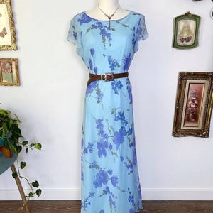 Op de afbeelding: Lichtblauwe maxi-jurk met bloemenprint, korte mouwen en een bruine riem. De jurk heeft een ronde hals en een zwierige rok. Het bloemenpatroon is in blauw- en paarstinten.