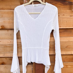 Puede incluir: Una blusa blanca de manga larga con textura arrugada, que presenta un escote en V, un cuerpo ajustado y mangas acampanadas que terminan en dobladillos puntiagudos e irregulares. La etiqueta interior del cuello dice DRESS U.