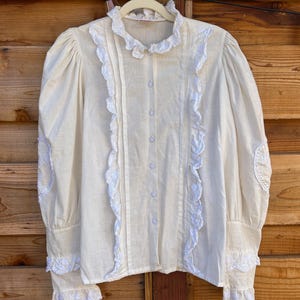 May include: A cream-colored long-sleeve blouse featuring a high ruffled collar, vertical white eyelet lace trim along the button placket, and matching eyelet lace detailing on the cuffs and elbows. The shirt has a button-down front closure.