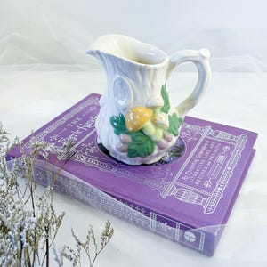 May include: A white ceramic creamer, shaped like a tree trunk, decorated with mushrooms and leaves. It rests on a purple book titled "The Faerie Handbook". The book features silver decorative elements.