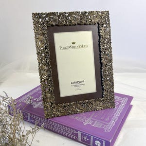 May include: An antique-style gold-plated picture frame with a dark brown interior, placed on a purple book. The frame has detailed scrollwork and the text "PHILIP WHITNEY LTD." The book is titled "THE FAERIE HANDBOOK."