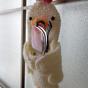 May include: A handmade, white crochet chicken-shaped eyeglass holder with a red comb and yellow feet. The chicken is holding a pair of spectacles. The holder is hanging on a window frame.