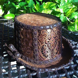 May include: A brown top hat with a gold and brown paisley pattern and a brown leather trim. The hat is sitting on a black metal surface.