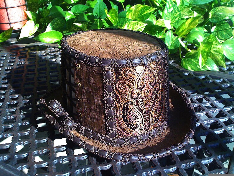May include: A brown top hat with a gold and brown paisley pattern and a brown leather trim. The hat is sitting on a black metal surface.