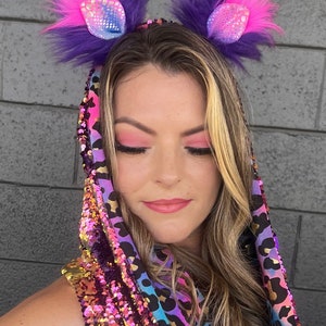 May include: A woman wearing a sequined hooded top with a leopard print design. She is wearing a pair of purple and pink furry cat ears with iridescent accents.
