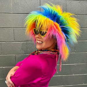 May include: A person wearing a bright, multi-colored wig with yellow, blue, pink, and red streaks. They are wearing sunglasses and a pink shirt.