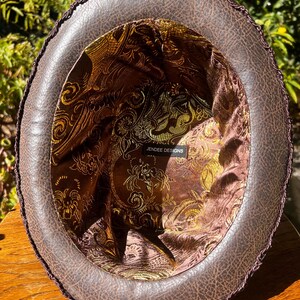 May include: The inside of a brown leather hat with a gold and brown patterned lining. The hat has a black trim around the edge. The label inside the hat reads "Jendree Designs".