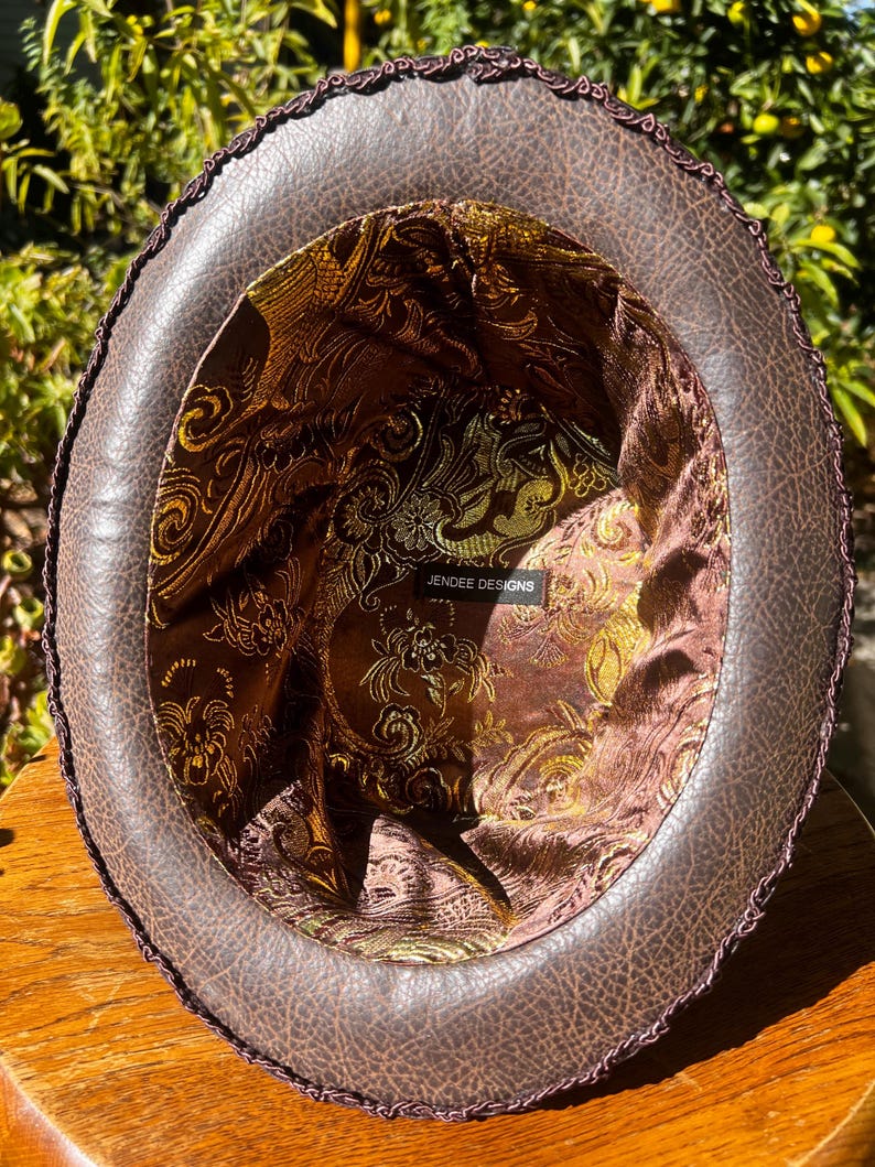 May include: The inside of a brown leather hat with a gold and brown patterned lining. The hat has a black trim around the edge. The label inside the hat reads "Jendree Designs".