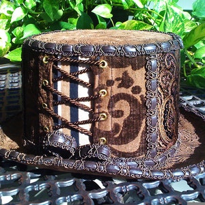 May include: A brown top hat with a brown and black patterned fabric and a brown leather trim. The hat has a corset-style lacing detail with brown cords and gold eyelets.