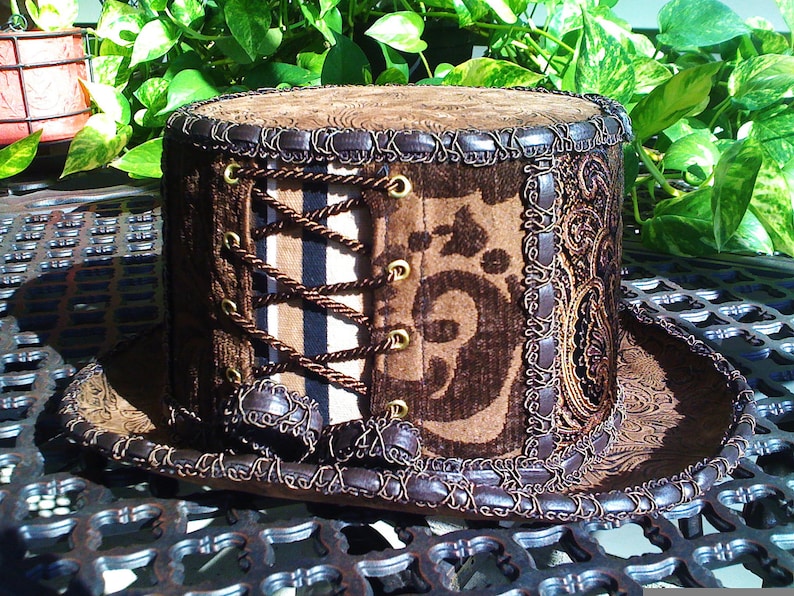 May include: A brown top hat with a brown and black patterned fabric and a brown leather trim. The hat has a corset-style lacing detail with brown cords and gold eyelets.