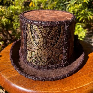 May include: A brown top hat with a paisley patterned band and a brown leather brim. The hat has a decorative stitched trim around the brim.