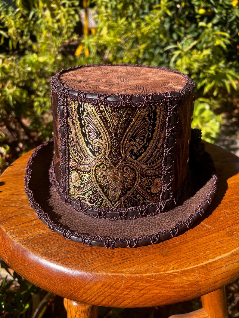 May include: A brown top hat with a paisley patterned band and a brown leather brim. The hat has a decorative stitched trim around the brim.