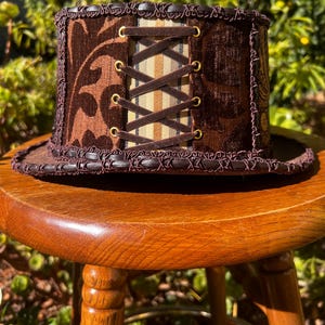 May include: A brown top hat with a brown and white patterned fabric band and a brown velvet top. The hat has a lace trim and a brown leather strap with brass eyelets.