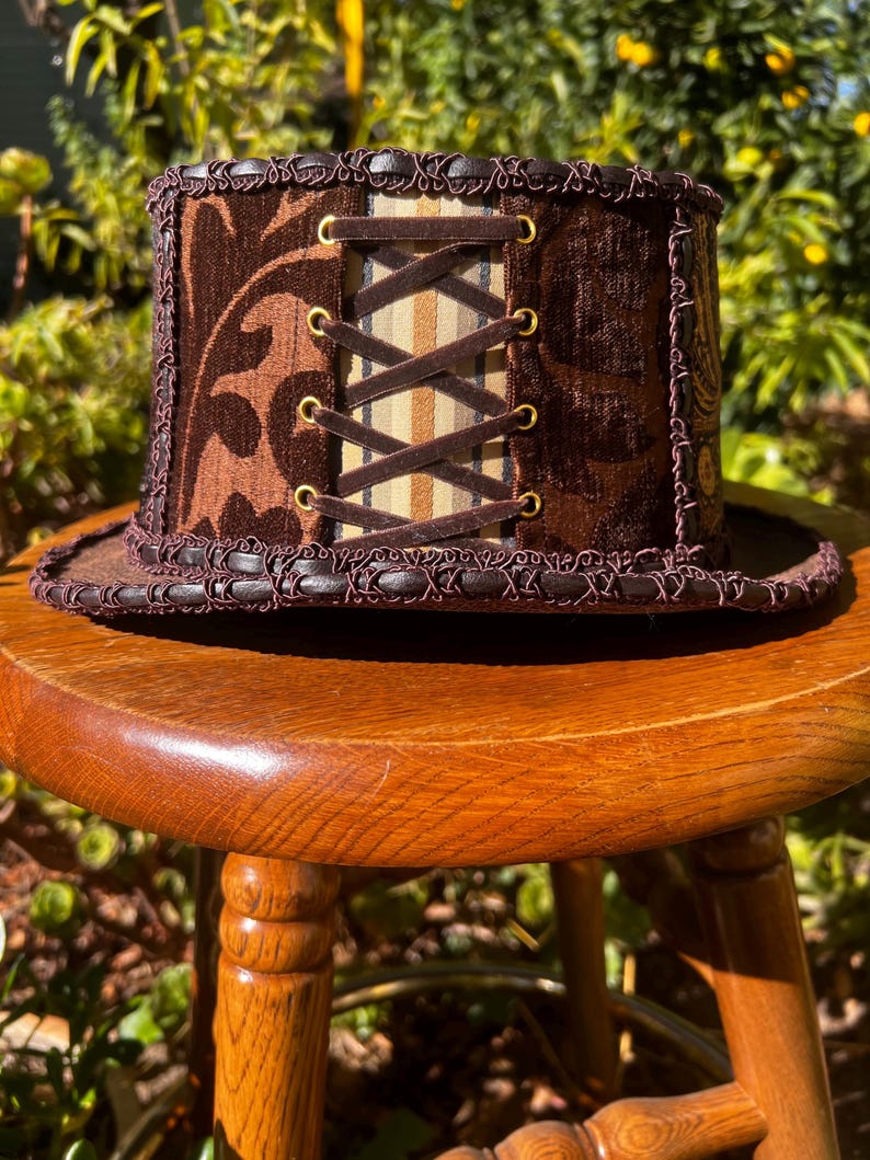 May include: A brown top hat with a brown and white patterned fabric band and a brown velvet top. The hat has a lace trim and a brown leather strap with brass eyelets.