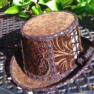 May include: A brown and gold top hat with a patterned design and a brown leather band. The hat has a lace-up detail on the side.